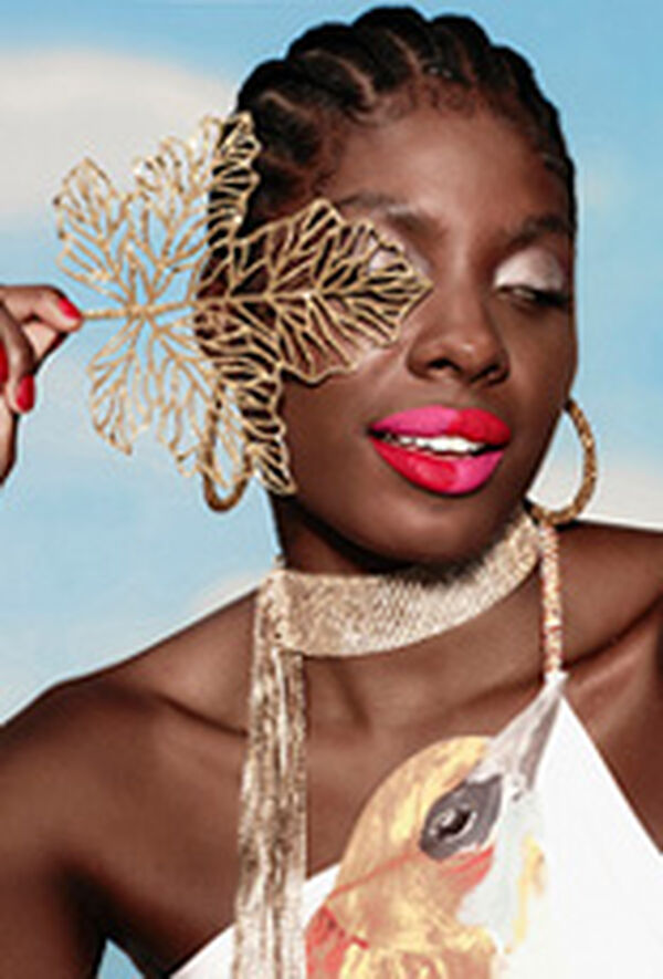 Mulher negra de cabelo trançado, com batom meio a meio nas cores rosa e vermelho. Ela segura um acessório dourado de folha diante do rosto e usa blusa branca com estampa de pássaro. Fundo azul.