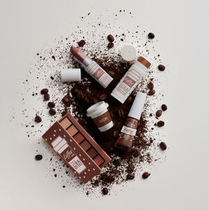 Batom e Multi Stick da linha Avon Color Trend Coffee Date mergulhando em xícara branca com conteúdo marrom leitoso. Fundo branco.