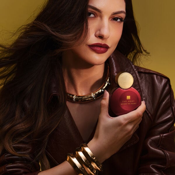 Imagem com mulher, branca, de cabelo ondulado longo e escuro e olhar intenso. Veste um casaco marrom dramático e joias douradas, e em uma das mãos segura o novo Deo Parfum Far Away Beyond The Secret.