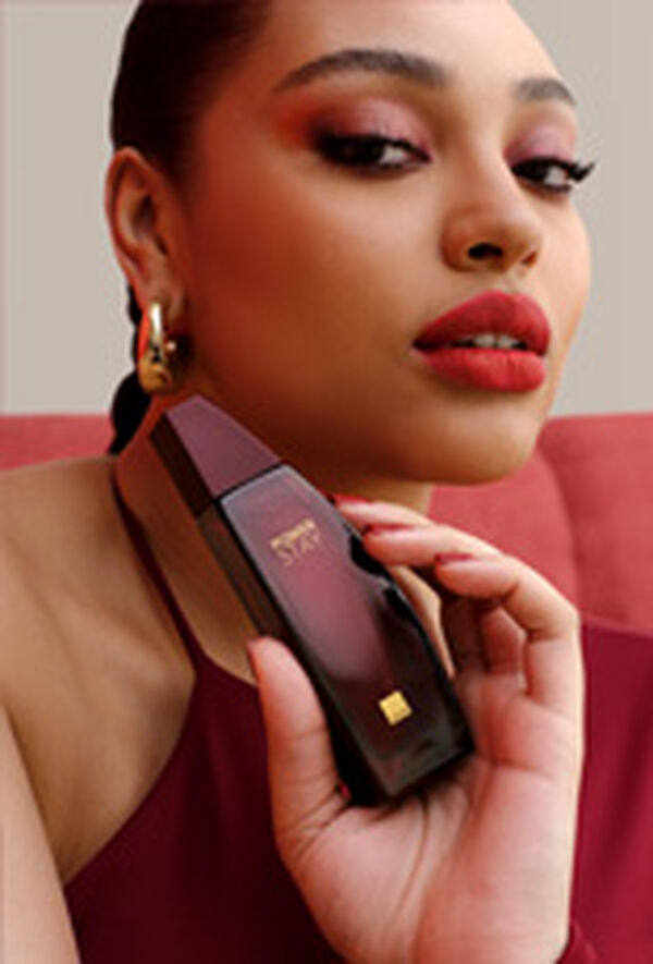 Mulher com maquiagem em tons de vermelho segura frasco de fragrância Power Stay em vermelho próximo ao  rosto. Ela usa vermelho e brincos dourados e está sentada em sofá vermelho em fundo claro.