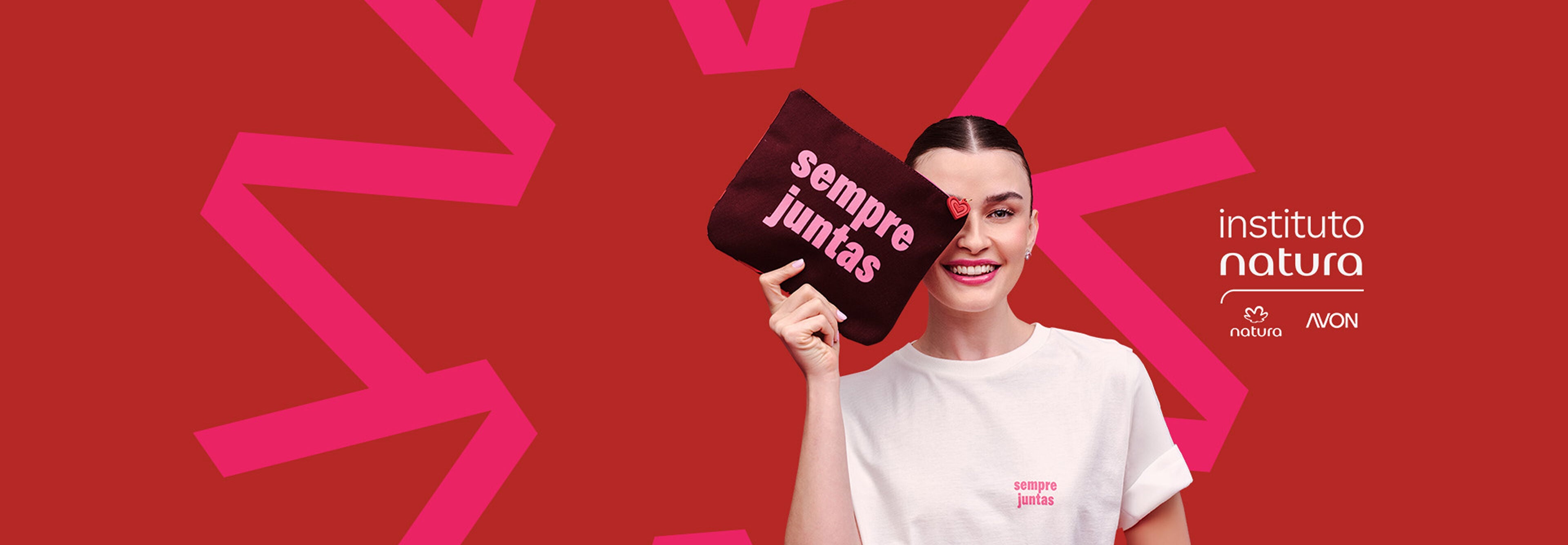 Mulher de pele clara sorri cobrindo um olho com necessaire escura escrito "sempre juntas". Ela veste camiseta branca e está diante de fundo vermelho com grafismos e marcas Instituto Natura e Avon.