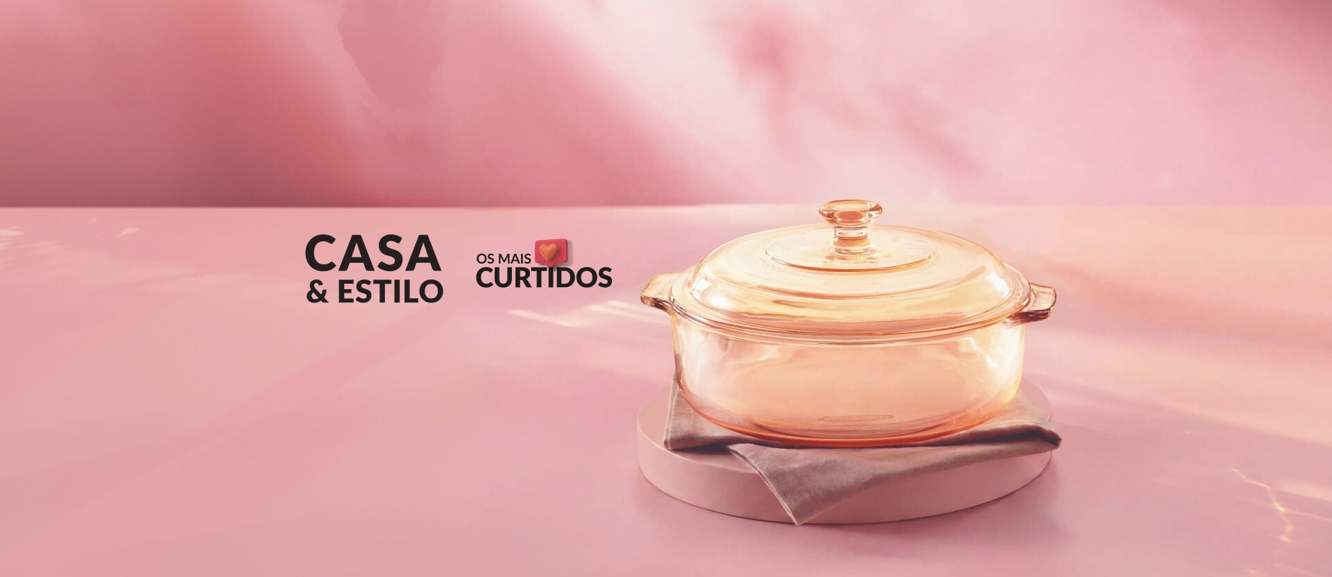 Banner promocional em ambiente rosa e iluminado. À direita, travessa refratária de vidro com tampa sobre guardanapo de tecido e suporte circular. À esquerda, o texto Casa & Estilo Os Mais Curtidos.