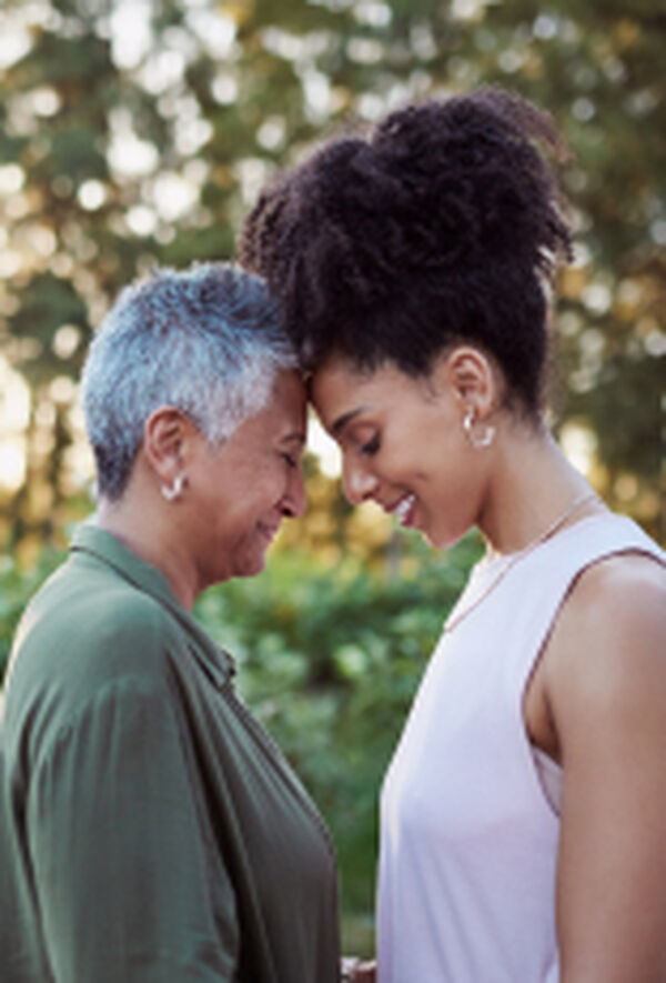 Mulheres negras de idades diferentes, frente a frente com as testas encostadas. À esquerda, mulher de cabelos curtos grisalhos de verde e, à direita, mulher de cabelos presos e crespos de rosa. Fundo externo doméstico.