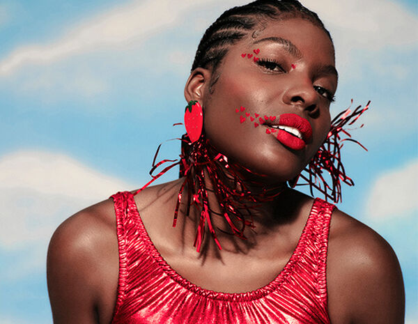Mulher negra de cabelo trançado, usando batom vermelho, brincos de morango e blusa vermelha brilhante. Sua maquiagem em tons de vermelho traz corações nos olhos e ao redor dos lábios. Fundo azul.