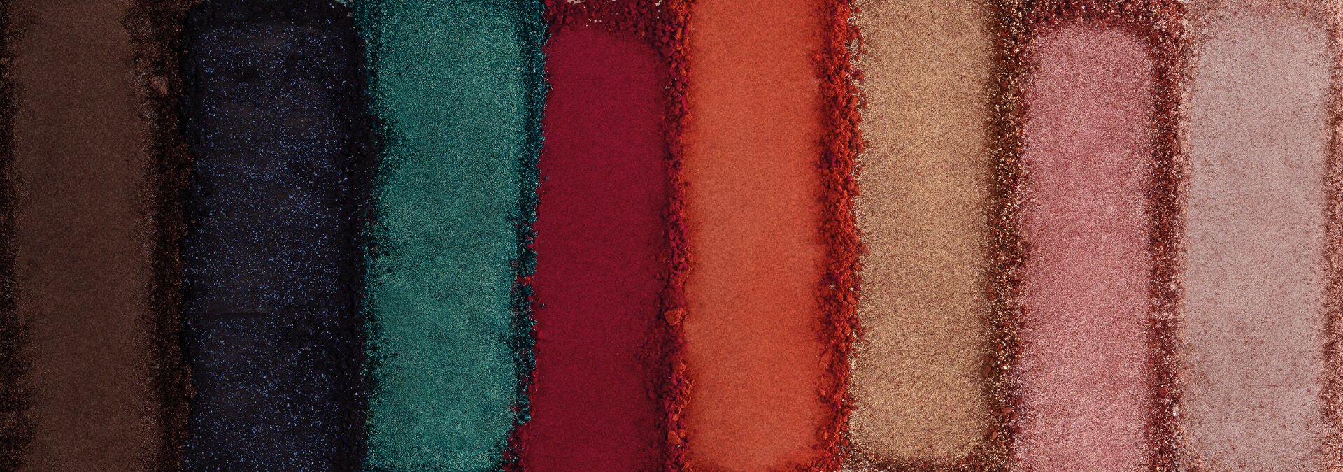 Banner com fundo bege claro. Elementos incluem lápis delineadores Avon em tons vibrantes de rosa e roxo, com traços desenhados ao redor. Texto: "Olhos: mais presentes, mais emoção."