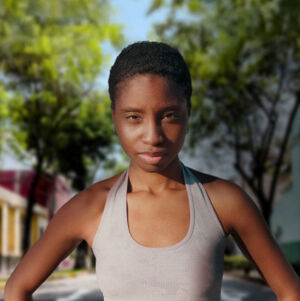 Foto de mulher negra de cabelos curtos e regata branca olhando para a câmera. O fundo é a imagem desfocada de uma rua arborizada.