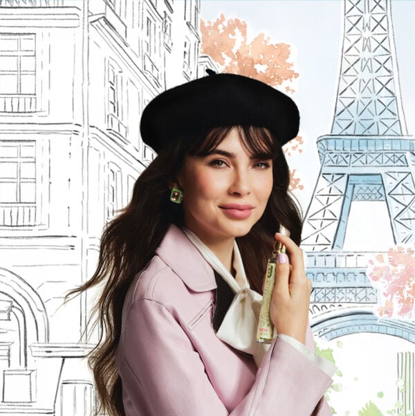 Imagem com fundo desenhado de Paris e em destaque uma mulher branca, de cabelo ondulado e escuro com franja, usando boina e blazer rosa. Ela segura próximo ao pescoço o Deo Parfum Macaron Amour.