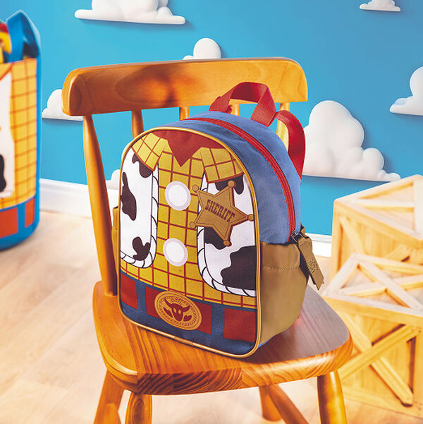 Cenário com parede azul e nuvens destaca uma mochila infantil estampada com a roupa do xerife Woody de Toy Story, apoiada sobre uma cadeira de madeira. Ao lado, há caixotes de madeira.