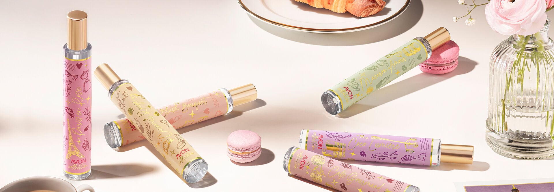 Imagem com os seis Deos Parfums da linha Avon Collection dispostos em uma superfície branca. A cena é composta com macarons, xícara de café, croissant e cartões-postais de Paris.