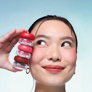 Mulher asiática com sorriso leve segura embalagens Avon Balm Multifuncional K-Jelly, K-Sorbet e K-Pudim. Faixa azul-claro.