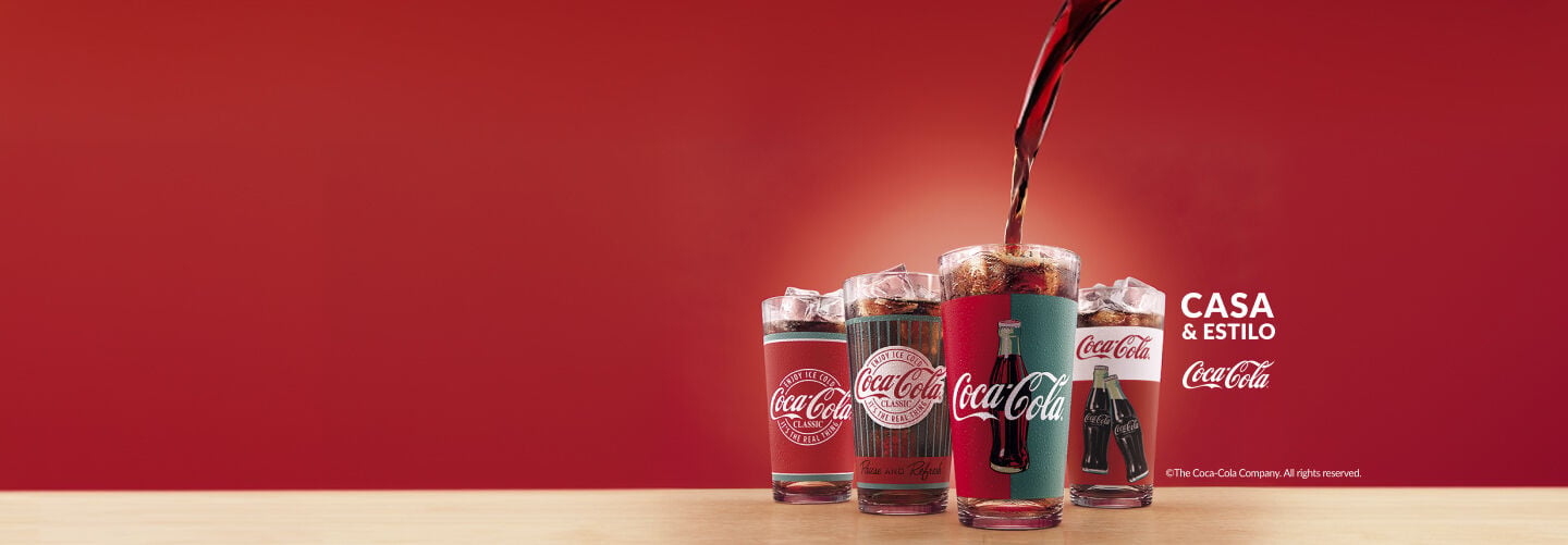 Quatro copos de vidro com gelo e estampas vintage da Coca-Cola alinhados sobre bancada de madeira, com o refrigerante sendo servido em um deles sob fundo vermelho vibrante.