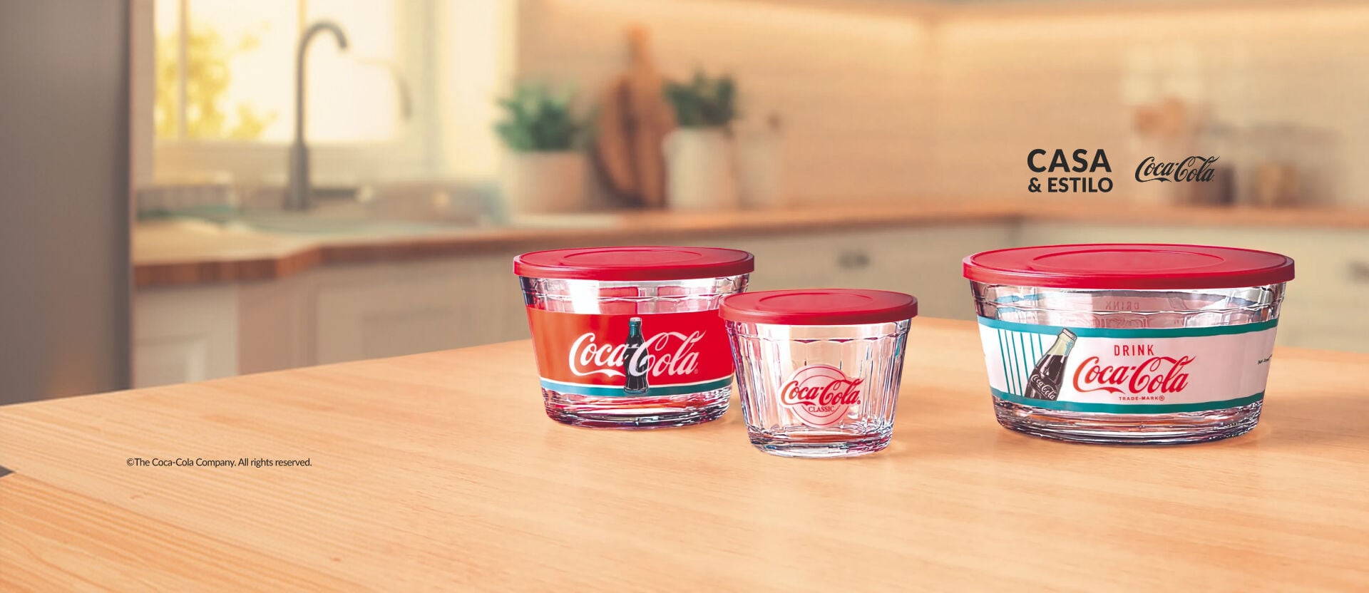 Três potes de vidro redondos com tampa plástica vermelha de tamanhos variados, decorados com estampas temáticas e o logotipo clássico da Coca-Cola, organizados sobre balcão de cozinha iluminado.