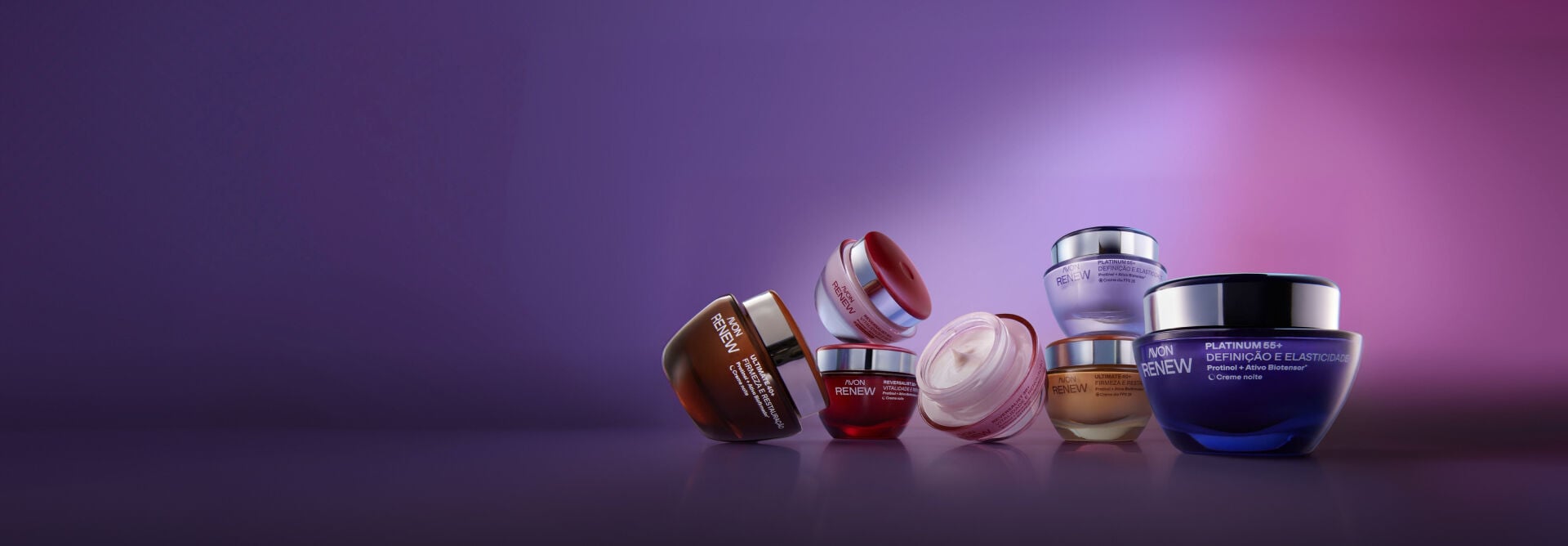 Linha Avon Renew reunida sob fundo roxo iluminado. Exibe potes de cremes faciais em tons de vermelho, dourado e azul, um deles aberto mostrando a textura cremosa do produto.