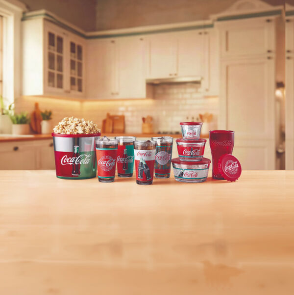 Conjunto completo de produtos da parceria Casa & Estilo e Coca-Cola dispostos sobre bancada amadeirada: balde de pipoca, diversos copos de vidro estampados, potes de mantimentos e copo vermelho. 
