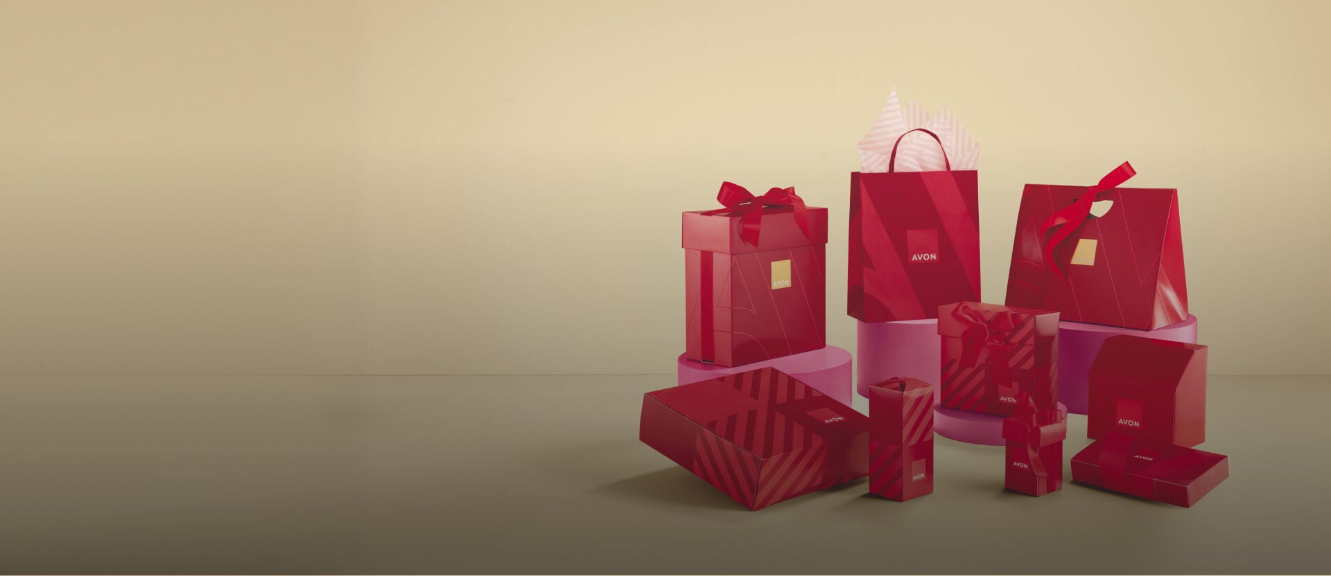Diferentes embalagens de presente vermelhas da Avon, incluindo caixas e sacolas, com o logo Avon em destaque. Elas estão dispostas sobre plataformas rosa. Fundo bege.