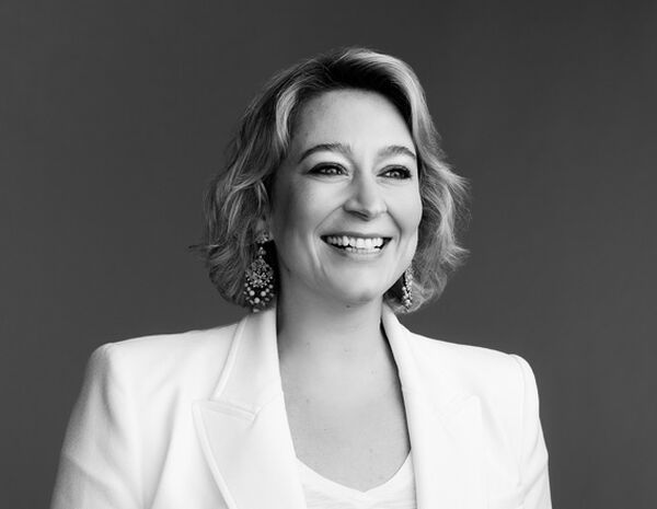 Foto em preto e branco de mulher sorrindo com cabelos loiros médios, usando roupa branca e brincos. 