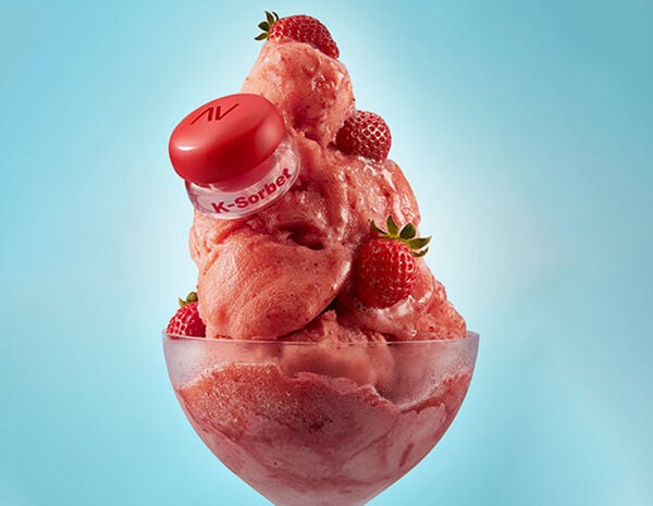 Embalagem Avon Balm Multifuncional K-Sorbet sobre sorvete com pedaços de morangos em taça translúcida. Fundo azul-claro.