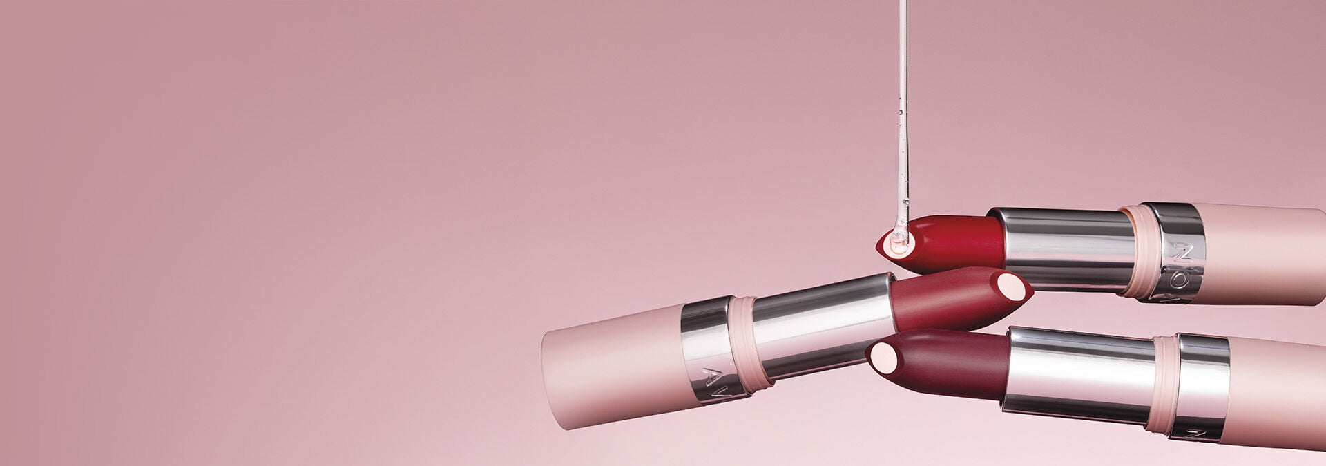 Banner com fundo rosa suave, apresentando batons em diferentes tons com embalagens elegantes. O texto destaca "Batom" e sugere hidratação e pigmentação intensa para um acabamento perfeito.