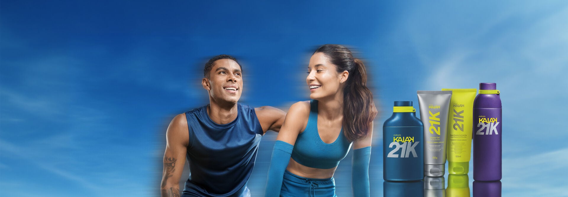 Banner sob céu azul com homem e mulher sorridentes em trajes de treino. À direita, frasco azul, shower gel, deo creme atrito e perfume roxo da linha Natura Kaiak 21K.