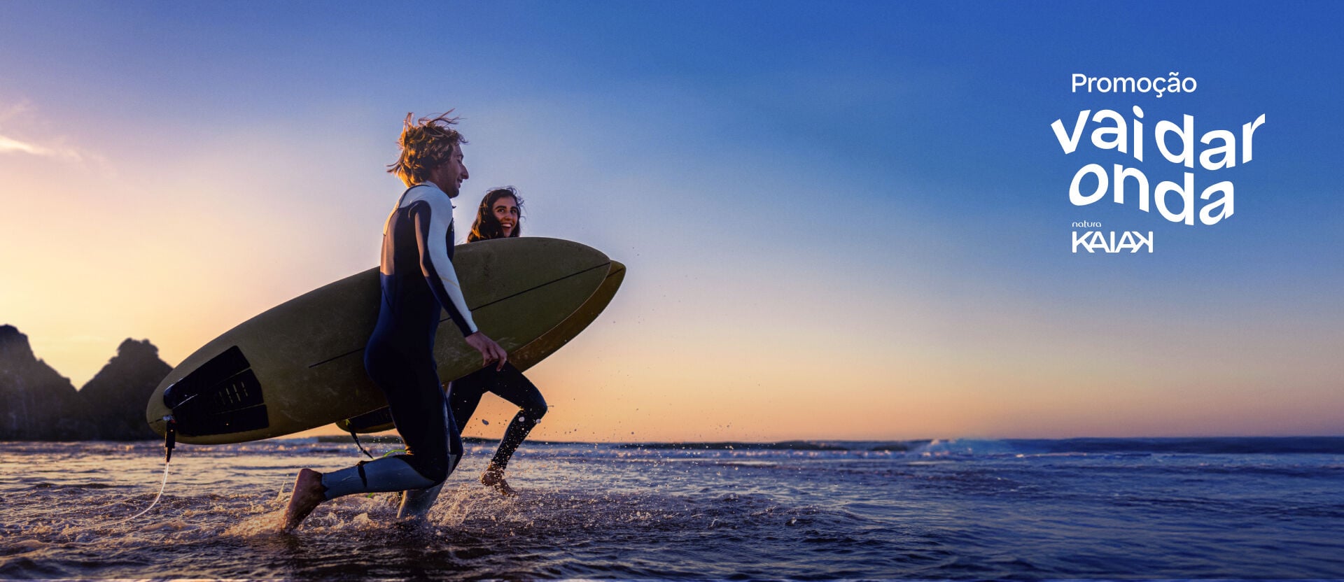 Homem e Mulher de pele branca, com macacões e pranchas de surfe, caminham em direção ao mar com a paisagem de Fernando de Noronha ao fundo. Selo aplicado: Promoção Vai Dar Onda Natura Kaiak.