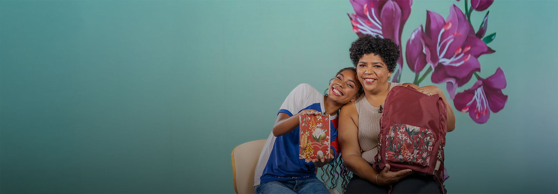 Uma adolescente e uma mulher negras sentadas juntas, sorrindo e mostrando uma caixa de presente e uma mochila com estampas florais da linha Natura Crer Para Ver.