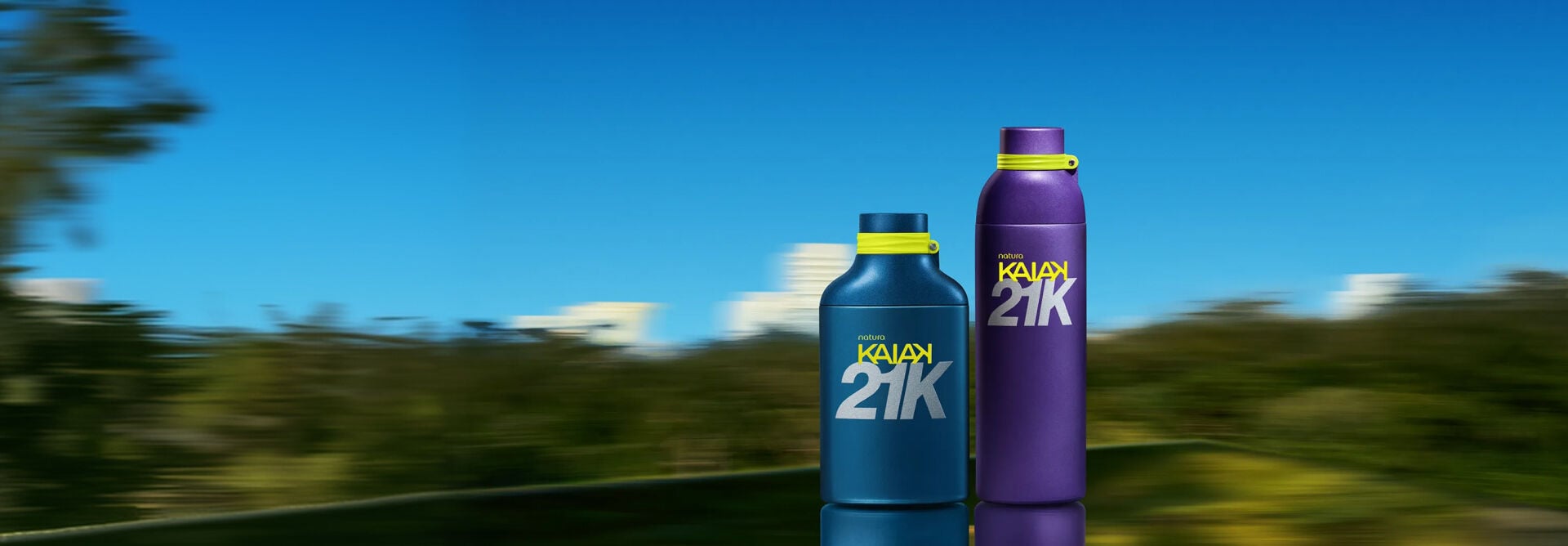 Imagem em fundo que remete a um parque com lagoa e céu azul, embaçado como uma captura de foto em movimento. Em destaque o Desodorante Colônia Kaiak 21K Masculino e o Deo Colônia Kaiak 21K Feminino.