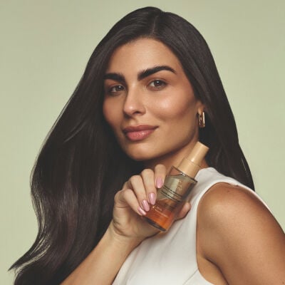 Mulher de pele morena, cabelos castanhos longos e blusa branca em fundo bege-claro. Ela segura o Óleo Bifásico Lumina Nutrição de Nanoprecisão, com frasco transparente de base laranja e tampa dourada.