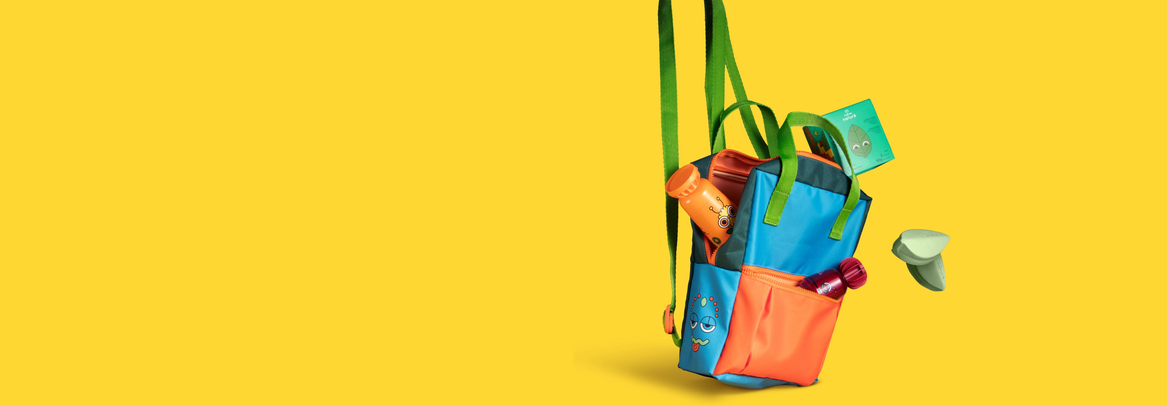 Mochila infantil colorida em fundo amarelo com produtos Natura Naturé saindo do zíper e do bolso. Entre os itens, estão: shampoo 2 em 1, colônia e sabonetes em barra.