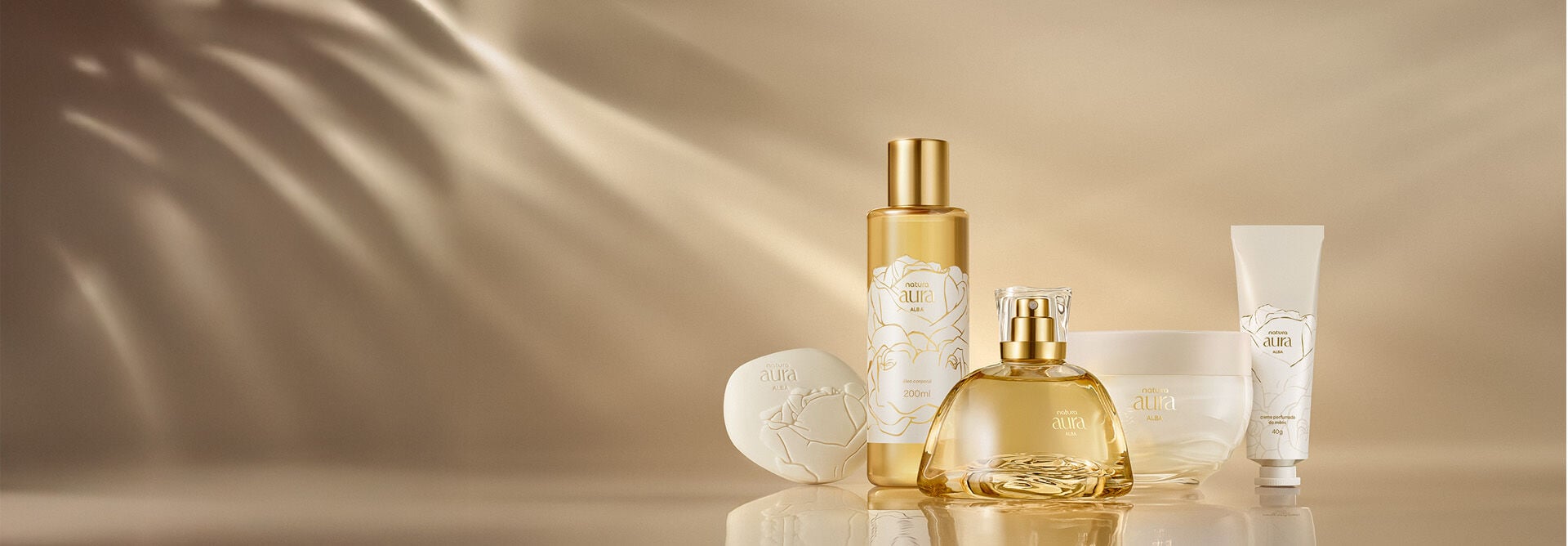 Linha completa de Natura Aura Alba, incluindo Sabonete Perfumado em Barra, Óleo Perfumado Corporal, Deo Parfum, Creme Corporal e Creme Perfumado de Mãos. Fundo dourado com reflexos.