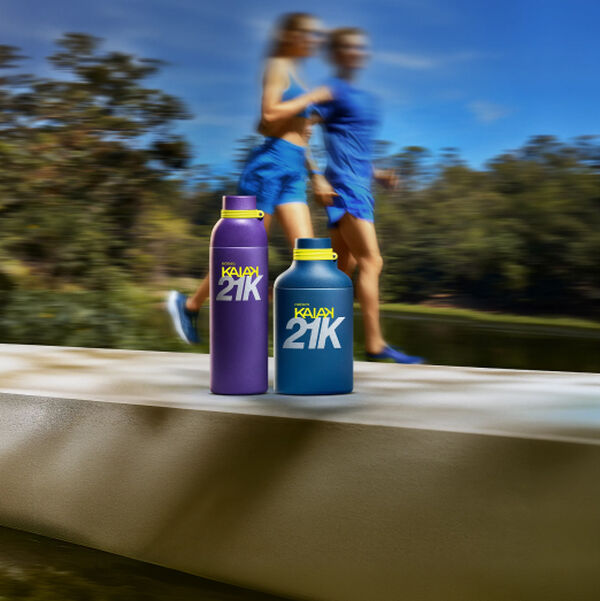 Banner sob céu azul com homem e mulher sorridentes em trajes de treino. À direita, frasco azul, shower gel, deo creme atrito e perfume roxo da linha Natura Kaiak 21K.