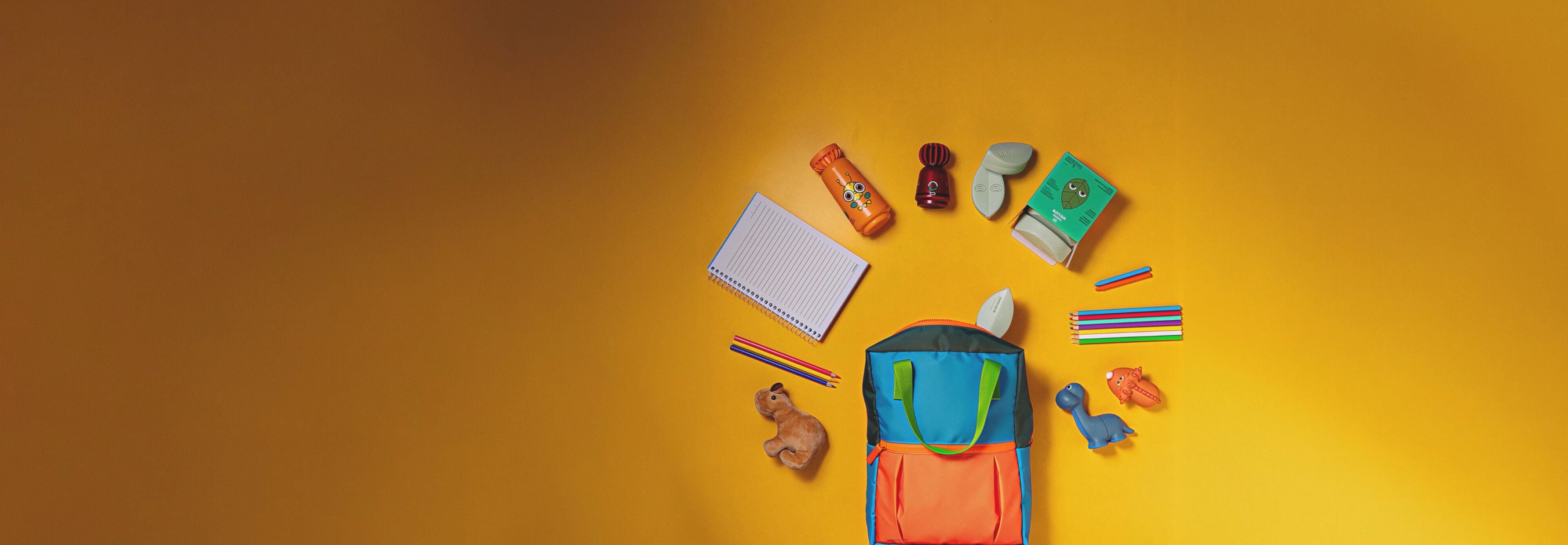 Mochila infantil colorida em fundo amarelo com produtos Natura Naturé saindo do zíper e do bolso. Entre os itens, estão: shampoo 2 em 1, colônia e sabonetes em barra.