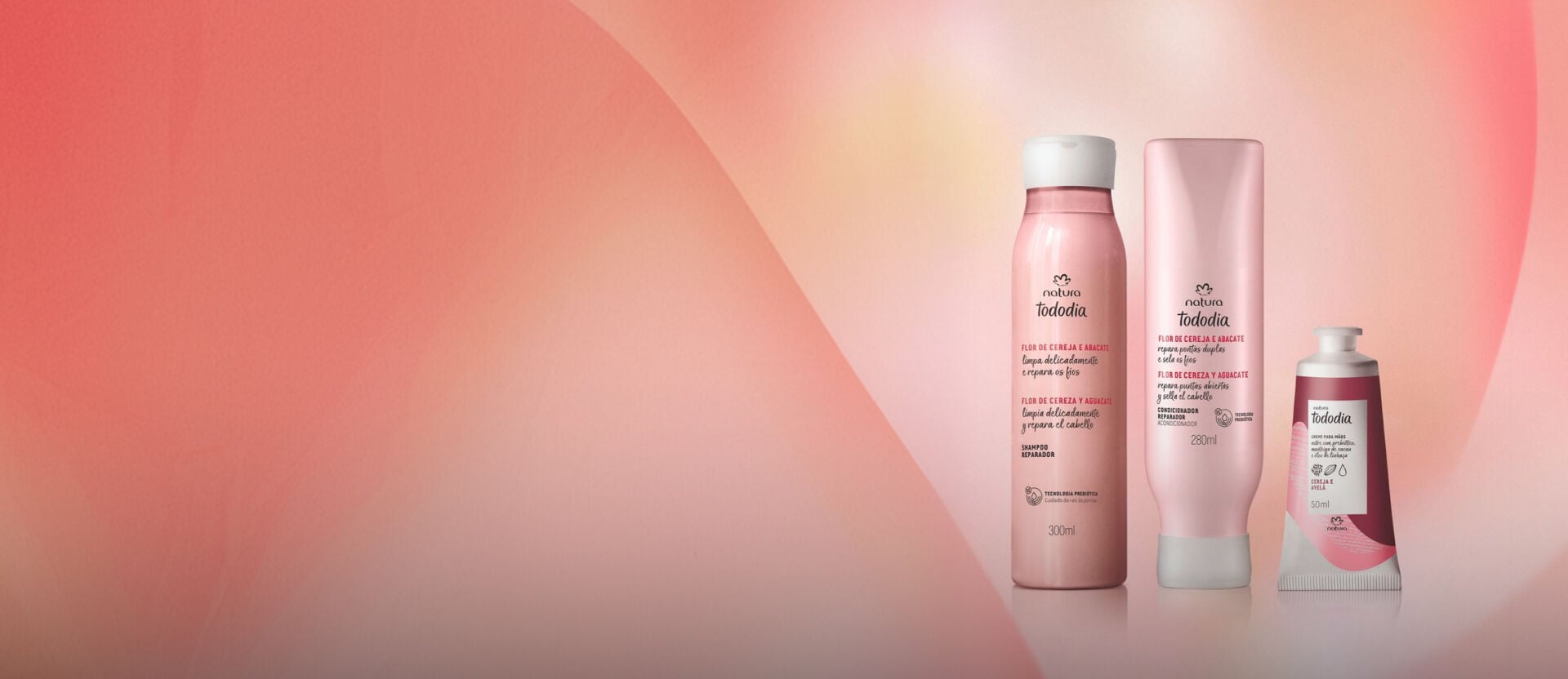 Shampoo e Condicionador Tododia Flor de Cereja e Abacate, com frascos rosa-claro e tampas brancas, ao lado do Creme para Mãos Cereja a Avelã, com bisnaga em tons de vermelho e rosa. Fundo rosado.