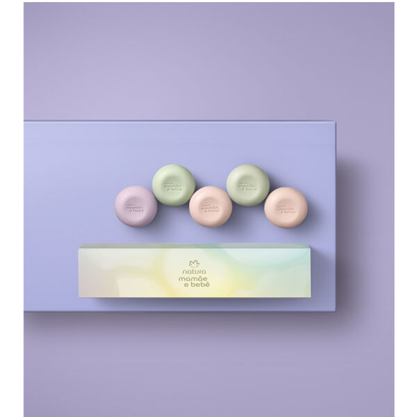 Cinco sabonetes redondos Natura Mamãe e Bebê em tons pastéis de verde, lilás e bege organizados sobre base lilás. Logo abaixo, a caixa retangular em tom suave exibe a marca.