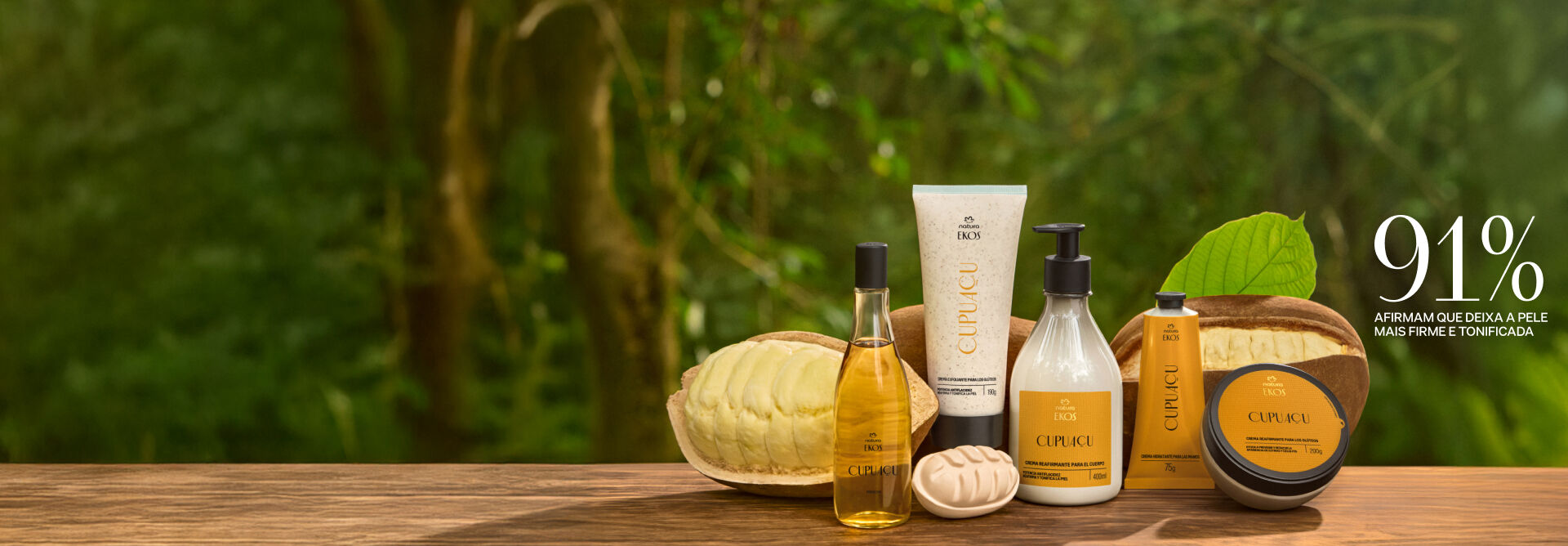 6 produtos da linha Ekos Cupuaçu, incluindo frescor, esfoliante, sabonete e cremes para corpo e mãos. Embalagens com rótulos amarelos sobre superfície de madeira, junto de frutos inteiros e cortados, em um fundo desfocado de folhagens. Texto: 91% afirmam que deixa a pele mais firme e tonificada.