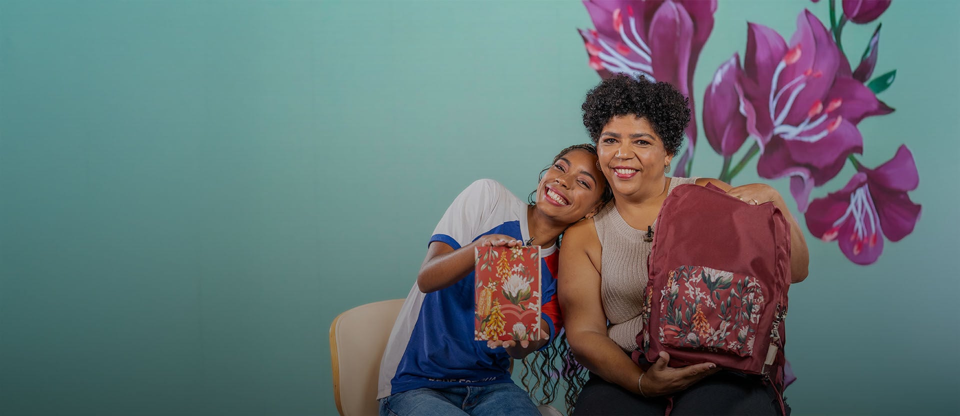 Uma adolescente e uma mulher negras sentadas juntas, sorrindo e mostrando uma caixa de presente e uma mochila com estampas florais da linha Natura Crer Para Ver.