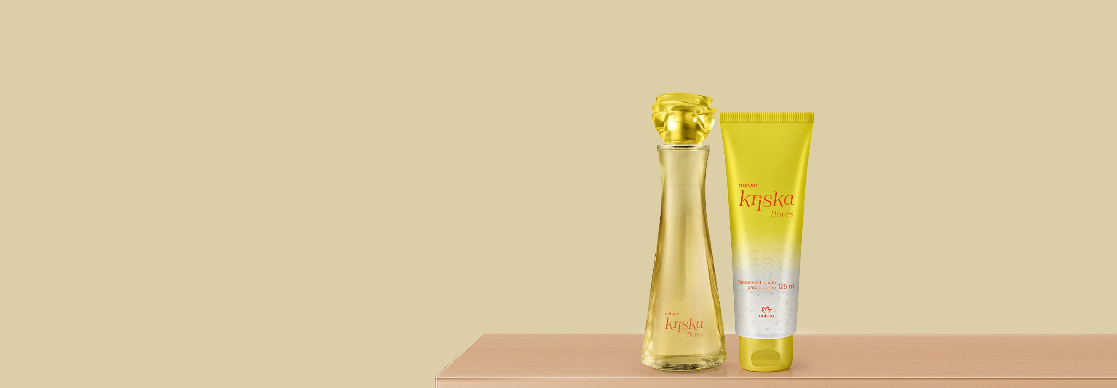 Frasco de perfume e bisnaga de sabonete líquido Natura Kriska Flores alinhados sobre bancada. As embalagens amarelas e translúcidas destacam o tom dourado sobre fundo neutro.