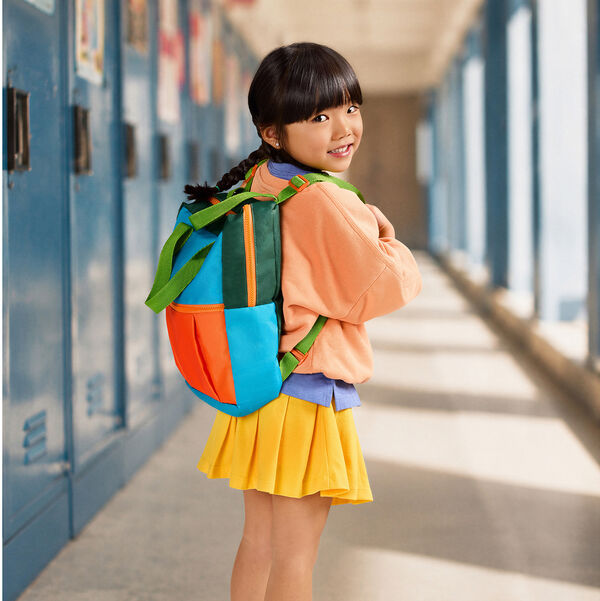 Menina sorri no corredor da escola olhando para trás. Ela veste blusa salmão, saia amarela e carrega nas costas a mochila infantil Natura Naturé colorida em tons de azul, verde e laranja. 