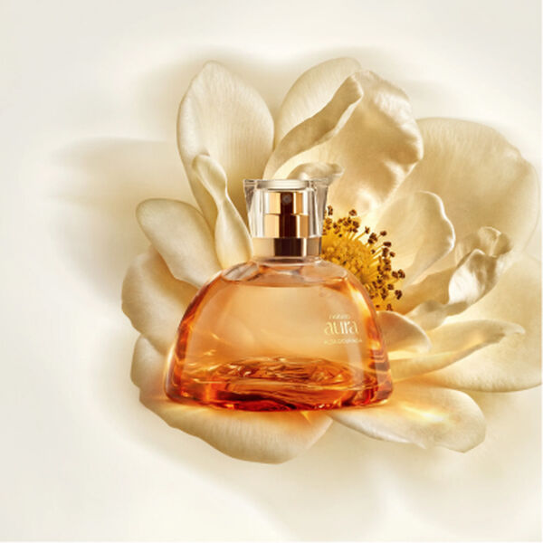 Imagem do novo Eau de Parfum Natura Aura Alba Dourada, um frasco translúcido luxuoso com líquido dourado, com uma Flor de Mel ao fundo.