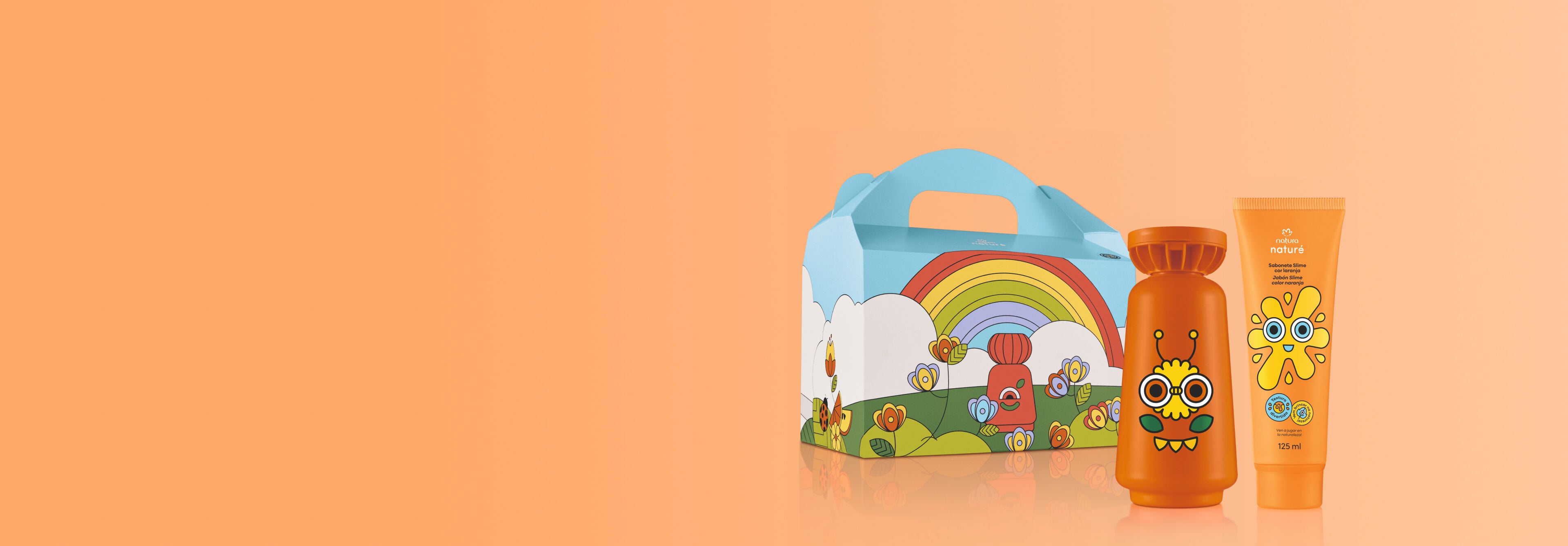 Kit Natura Naturé sobre fundo salmão com caixa de presente ilustrada com arco-íris e flores. Ao lado, frasco de colônia e bisnaga de sabonete slime laranjas trazem rostos divertidos.
