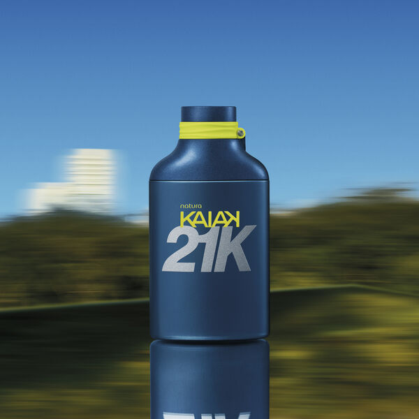 Frasco azul-escuro fosco de perfume Natura Kaiak 21K com letras prateadas e detalhe amarelo na tampa, contra fundo de natureza com efeito de velocidade. 