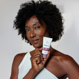 Mulher negra segura Multiprotetor FPS 70 próximo ao rosto. Ela usa blusa branca de ombro único e está em fundo branco