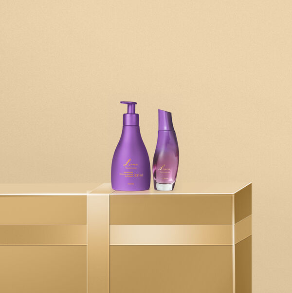 Frasco de perfume curvilíneo e hidratante corporal Natura Luna Fascinante roxos dispostos sobre suporte dourado. O fundo bege minimalista destaca o tom lilás elegante e o design das embalagens.