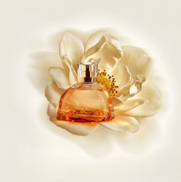 Frasco de perfume Natura Aura Alba Dourada em vidro translúcido ambarado repousa sobre uma grande flor branca aberta. O fundo claro em tom de creme destaca a sofisticação da embalagem.