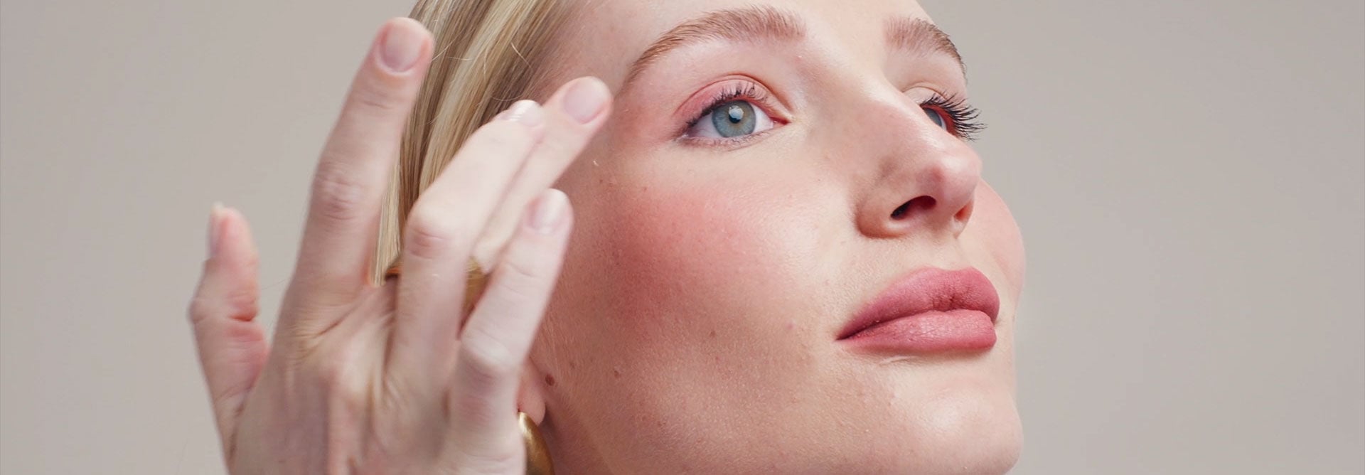Modelo de pele clara e cabelo loiro liso demonstra a versatilidade do Batom Blur Natura Una. Ela aplica o produto de textura aveludada nos lábios, esfuma como blush nas bochechas e usa como sombra nas pálpebras, destacando um acabamento suave em tom rosado. Ao longo do vídeo, frascos do batom em diferentes nuances são exibidos em destaque.