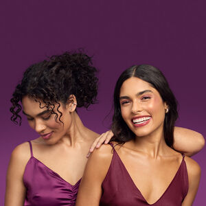 Foto de duas mulheres, uma de cabelos cacheados e presos e outra com os cabelos soltos, em fundo roxo. Ambas usam vestido de alças em tons de vinho.