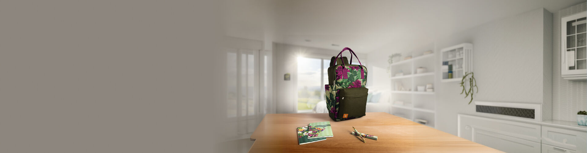 Mochila Natura Crer Para Ver com estampa floral verde e rosa sobre mesa de madeira ao lado de cadernos e canetas. O fundo exibe uma sala iluminada com estantes e janela.