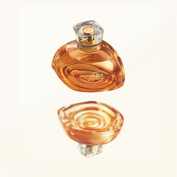 Frasco de perfume Natura Aura Alba Dourada em vidro translúcido ambarado repousa sobre uma grande flor branca aberta. O fundo claro em tom de creme destaca a sofisticação da embalagem.