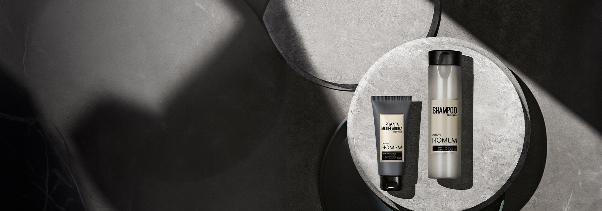 Fundo com tons de cinza com sombras e ao lado direito o shampoo 2 em 1 e a pomada modeladora de Natura Homem.