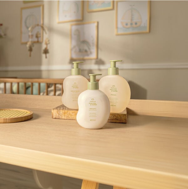 Quarto de bebê com berço e decoração ao fundo. Na bancada, as novas embalagens 400 ml dos produtos Mamãe e Bebê, sendo eles Sabonete Líquido, Hidratante e Shampoo.