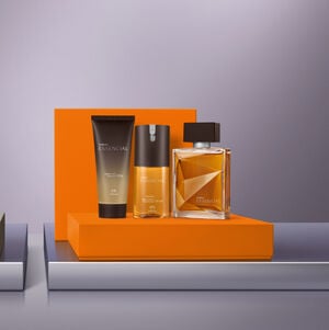 Kit de perfumaria masculina Natura Essencial sobre um bloco laranja, contendo uma bisnaga de shower gel, um desodorante spray e o perfume clássico em frasco de vidro geométrico com líquido âmbar.